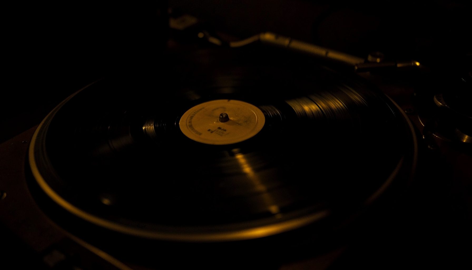 A dark, low-key photograph of a vinyl record spinning on a turntable, with deep shadows and warm amber highlights catching the grooves.