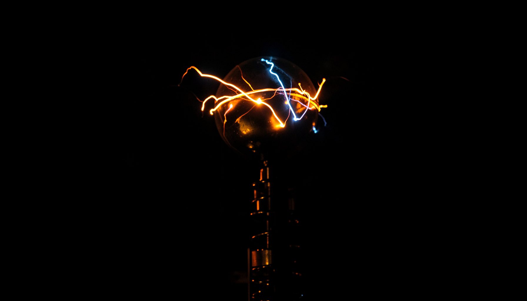 A dark, low-key close-up of glowing orange and blue electrical arcs dancing across a metal sphere atop a Tesla coil in near-total darkness.