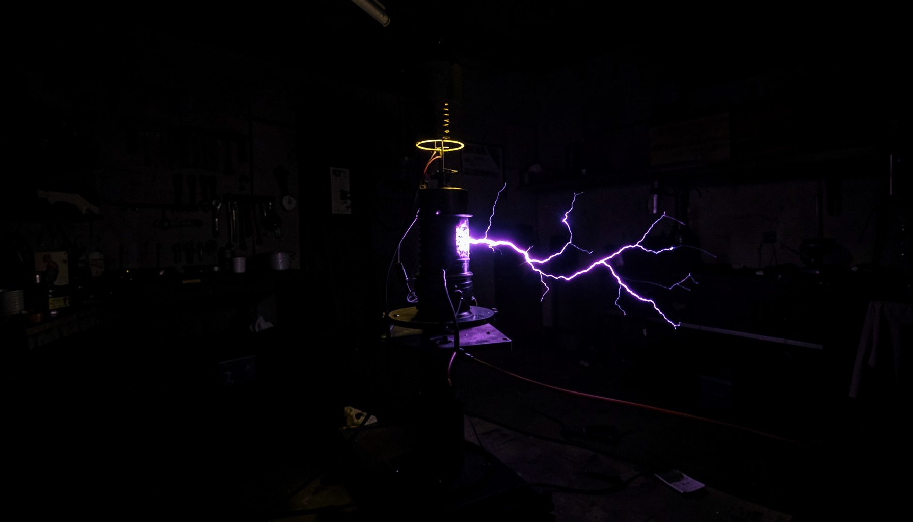 A dark, low-key photograph of a Tesla coil discharging bright violet-blue electrical arcs in a dim workshop, casting dramatic shadows.