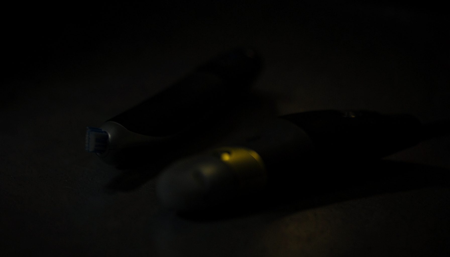 Dark close-up of two electric toothbrushes resting side by side on a matte surface, deep shadow and subtle highlights, moody and understated